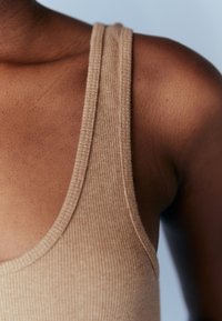 Gros plan sur l’épaule et le haut de la poitrine d’une personne portant un débardeur côtelé beige sur un fond clair.