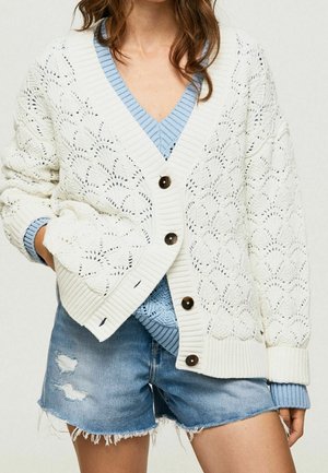 Bílý pletený cardigan s průstřihovým vzorem, žebrovanými manžetami a knoflíky. Nošený přes světle modrý svetr s výstřihem do V, doplněný džínovými šortkami.