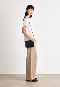 Borsa a tracolla nera con patta e accessori in argento, abbinata a una maglietta bianca con piping neri e pantaloni beige a wide-leg.