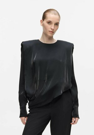 Žena s uhladenými vlasmi, ktorá má na sebe lesklý čierny oversize top s polstrovanými ramennami a čierne nohavice, stojí na jednoduchom bielom pozadí.