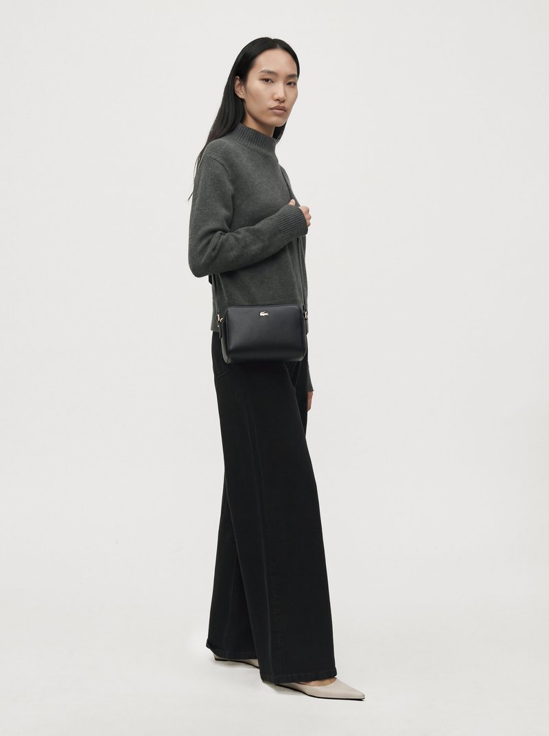 Une femme porte un pull anthracite, un pantalon noir à jambes larges et un sac bandoulière noir sur un fond clair et uni.