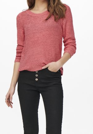 Pull en tricot rose clair avec un col rond et des manches longues, associé à un jean noir taille haute avec des boutons visibles.