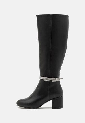 Schwarzer knöchelhoher Lederstiefel mit Blockabsatz und silbernem geflochtenem Riemen, der um den Knöchel auf einem weißen Hintergrund gebunden ist.