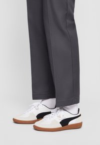 Białe i czarne sneakersy wykonane z zamszu i skóry, z brązową gumową podeszwą i białymi sznurowadłami, w zestawieniu z szarymi spodniami.