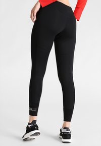 Donna che indossa leggings neri a vita alta e scarpe sportive nere con suole bianche, in piedi su uno sfondo chiaro e neutro.