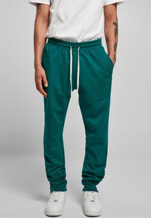 Teal joggebukser med elastisk midje og snor, med lommer på siden og en glatt tekstur, sammen med hvite joggesko.