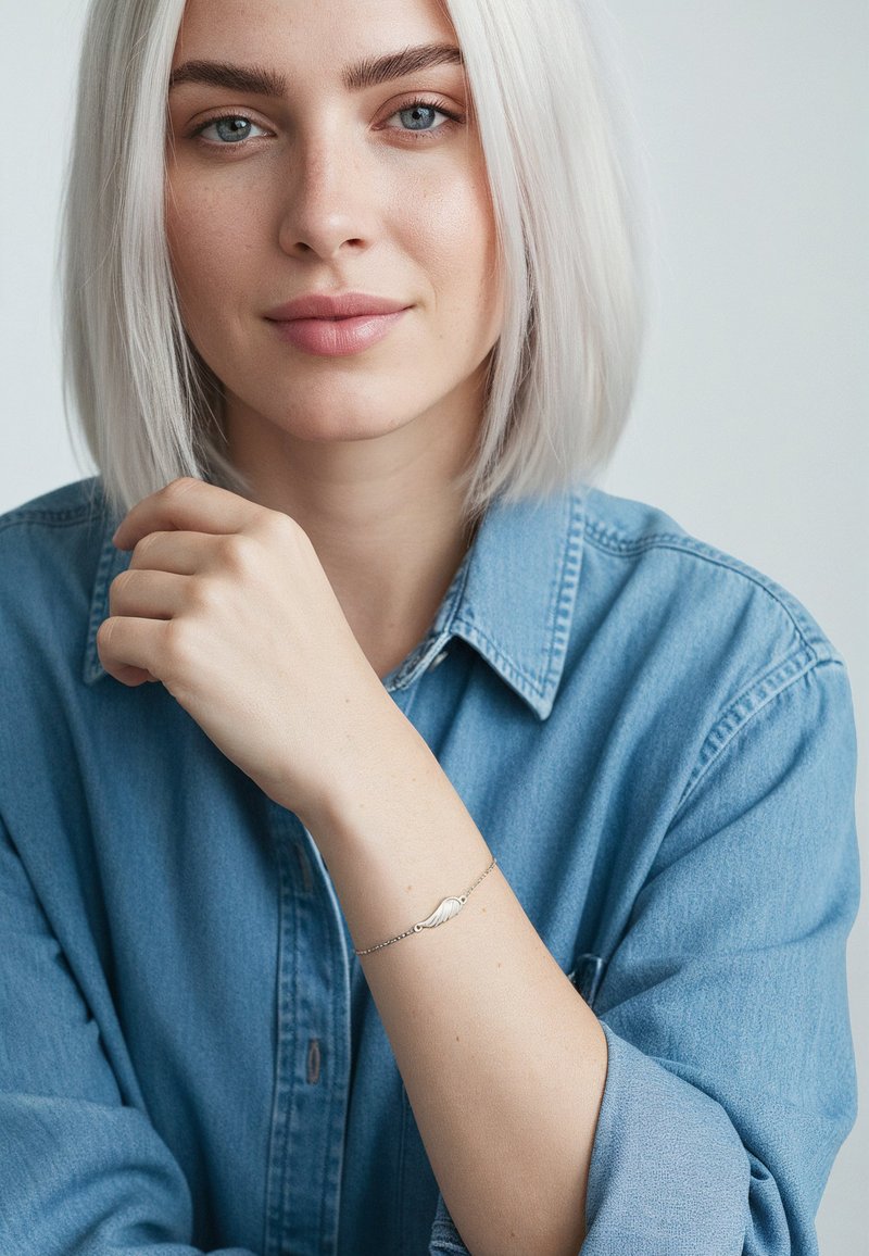Bracciale d'argento con dettaglio a forma di ala su una catena delicata, indossato su un polso nudo. Modello che indossa una camicia di jeans azzurro chiaro.