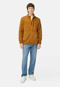 Orange tekstureret fleecejakke med høj krave, frontlynlås og sidelommer, parret med blå jeans og hvide sneakers.