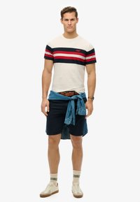 Camiseta de manga corta color crema con rayas azul marino y rojas, combinada con pantalones cortos azul marino. Camisa de mezclilla atada alrededor de la cintura, zapatillas color crema y calcetines estampados.