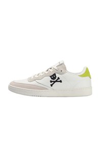 Zapatilla blanca de corte bajo con acentos de ante beige, logo de calavera y tibias negras en el lateral, y detalle amarillo brillante en el talón.