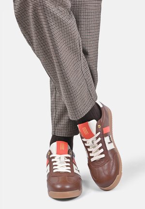 Persoon die bruine en rode leren sneakers met witte veters en geruite bruine broek draagt tegen een effen achtergrond.