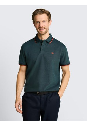 Mann mit dunkelgrünem Polohemd mit rotem Rand und marineblauen Hosen, der mit einer Hand in der Tasche vor einfarbigem Hintergrund lächelt.