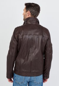 Bruine leren jas met een hoge kraag, gestikte schouderaccenten en zijzakken. Gladde textuur en gestructureerd ontwerp met zichtbare stiksels.