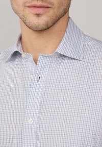 Primer plano de un hombre que lleva una camisa blanca de vestir con un patrón de cuadros azules y el cuello desabrochado, mostrando una leve barba de tres días.