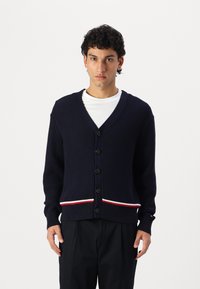 Marineblå strikk cardigan med v-hals, svarte knapper og rød-hvitt stripet kant. Teksturert stoff med lange ermer.