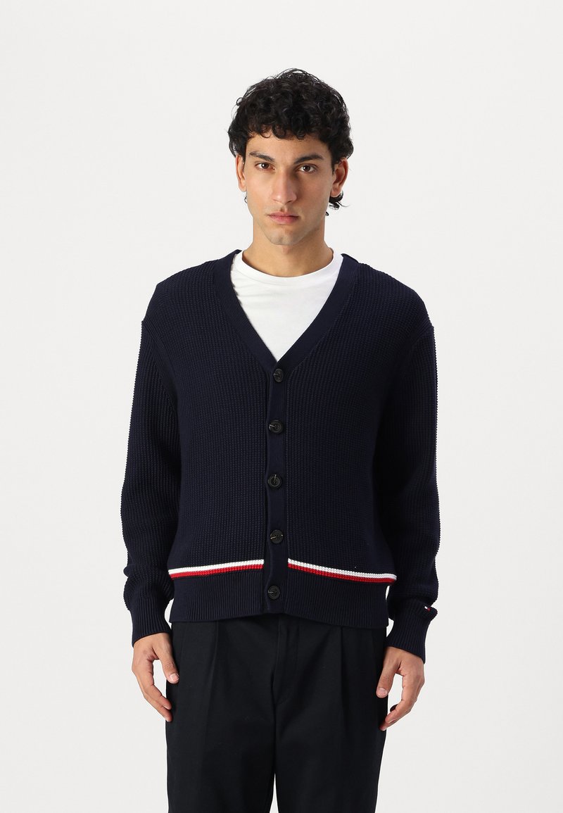 Marineblå strikk cardigan med v-hals, svarte knapper og rød-hvitt stripet kant. Teksturert stoff med lange ermer.