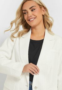 Weißer strukturierter Blazer mit einem Ein-Knopf-Verschluss, zwei frontalen Taschen und einem Reverskragen, getragen über einem schwarzen Hemd und blauen Jeans.