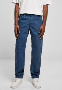 Pantalon en coton bleu foncé à coupe droite, avec poches, braguette zippée et détails de couture subtils, associé à des baskets blanches.
