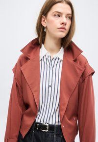 Νεαρή γυναίκα που φοράει σακάκι χρώματος σκουριάς πάνω από λευκό πουκάμισο με κάθετες μπλε ρίγες, συνδυασμένο με σκουρόχρωμα τζιν και ασημένια σκουλαρίκια κρίκους.