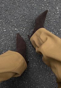 Chaussures à talons hauts en daim marron avec un bout pointu et un talon aiguille, assorties à un pantalon beige à jambes larges. Surface texturée visible.