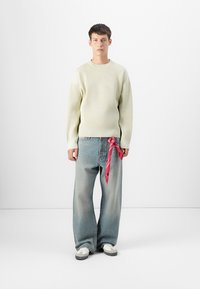 Pulover ribbed verde deschis, blugi lejeri albastre deschise, eșarfă roșie legată în talie, adidași albi, stând în fața unui fundal neutru.