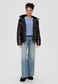 Czarna puchowa kurtka z kapturem, zapinana na zamek, z bocznymi kieszeniami, noszona na jasnoniebieskim swetrze z dzianiny i szerokich niebieskich dżinsach.