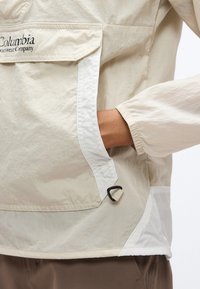 Άτομο που φοράει μπεζ αδιάβροχο μπουφάν Columbia, με το χέρι μέσα στην πλαϊνή τσέπη και ελαστικό μανσέτα στο μανίκι.