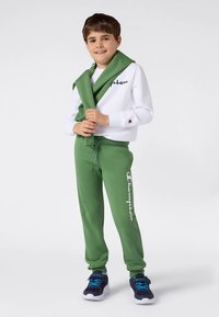 Weißes Sweatshirt mit Logo, grüne Sweatpants mit seitlichem Branding und blaue Sportschuhe. Accessoire: grüner Pullover locker über die Schultern gelegt.