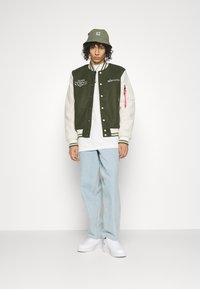 Giacca varsity verde e crema con dettagli ricamati, abbinata a una t-shirt bianca, jeans azzurri chiari e sneakers bianche. Cappello a secchiello verde.
