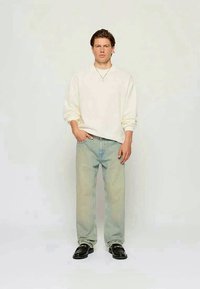 Cremefarbener Pullover mit überschnittenen Schultern, hellblaue Jeans und schwarze Mokassins, stehend vor einem neutralen Hintergrund, Hände in den Taschen.
