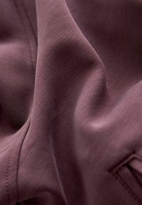 Gestructureerde stof in een gedempte mauve kleur, met een gladde, vloeiende uitstraling, subtiele stiksels en een klein logo-detail.