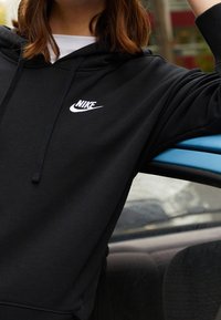 Zwarte hoodie gemaakt van zachte stof, met een wit Nike-logo op de borst. De mouwen zijn lang en hebben een ontspannen pasvorm.