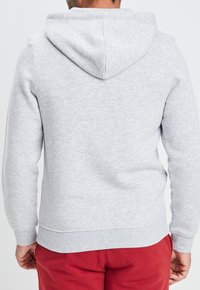 Sudadera gris con una tela suave y texturizada, con capucha y puños acanalados. Llevada con pantalones rojos, mostrando la vista trasera.