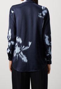 Chemise en satin bleu marine avec motif floral abstrait bleu clair, manches longues et ourlet arrondi. Texture lisse ; poignets boutonnés.