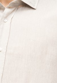 Chemise en lin beige clair avec un petit logo blanc brodé sur la poitrine, col et boutons visibles.