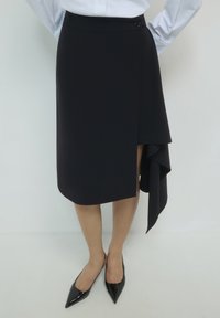 Donna che indossa una gonna nera asimmetrica con un dettaglio drappeggiato laterale, una blusa bianca e tacchi slingback neri a punta, in piedi con le mani dietro la schiena.