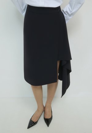 Frau in einem schwarzen asymmetrischen Rock mit seitlichem Drapierungsdetail, weißer Bluse und schwarzen spitzen Slingback-Absätzen, steht mit den Händen hinter dem Rücken.