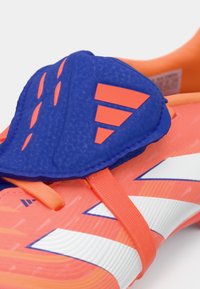 Zapato de fútbol naranja brillante con acentos azules y una cubierta protectora. Presenta patrones texturizados y varias franjas en blanco.