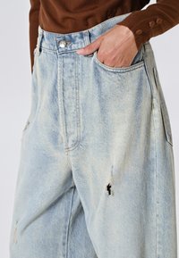 Jeans a gamba larga in denim blu chiaro con toppe strappate, vita alta, ampie tasche frontali e chiusura con bottone metallico.