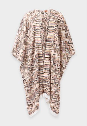 Une femme porte un sous-pull beige à col roulé, un cardigan à motifs ouvert sur le devant, un pantalon marron à jambes larges et des mocassins foncés sur un fond blanc uni.