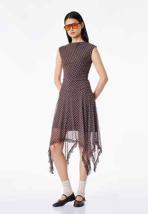 Kobieta ubrana w brązową sukienkę w kropki bez rękawów z asymetrycznym dołem, beżowe skarpetki, czarne buty typu Mary Jane oraz duże pomarańczowe okulary przeciwsłoneczne.