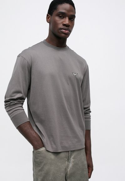 Garās piedurknes pelēks T-krekls, izgatavots no mīksta auduma, ar mazu logo kreisajā krūtī un ribota piedurkņu apdare.