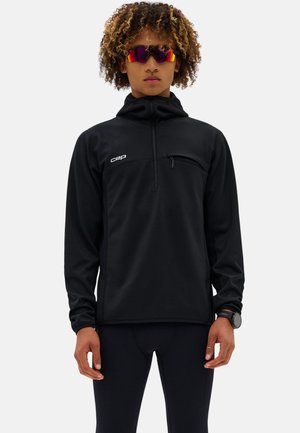 Persona con capelli ricci che indossa occhiali sportivi riflettenti, un pullover nero a maniche lunghe con zip e tasca sul petto, e pantaloni neri.