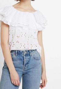 Vit off-shoulder blus med lager av volanger, prydd med blommiga hålskärningar. Kombinerad med högmidjade blå denimjeans.