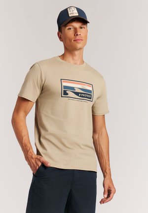 Beige katoenen T-shirt met een kleurrijke surfthema-afbeelding en korte mouwen, gecombineerd met een donkere short en een marineblauwe pet.