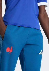 Modré športové tričko s bielym golierom, doplnené modrými nohavicami s červeným logom kohúta a bielymi pruhmi po boku a na logu.