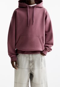 Persona che indossa una felpa ampia color rosa polveroso con tasca frontale e pantaloni patchwork a gamba larga beige chiaro, con le mani in tasca su uno sfondo bianco.