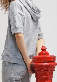 Sudadera gris con capucha y mangas cortas, de tejido texturizado, combinada con unos jeans gris claros. Hay un hidrante rojo en primer plano.