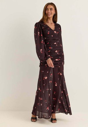 Kvinna står i en mörk, lång klänning med blommönster, långa puffärmar och svarta öppna klackskor mot en enfärgad ljus bakgrund.