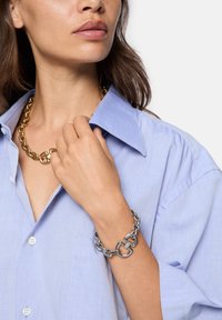 Collana in metallo dorato e braccialetto in argento indossati da un modello con una camicia colletto azzurro chiaro. Le catene sono spesse, con anelli interconnessi.